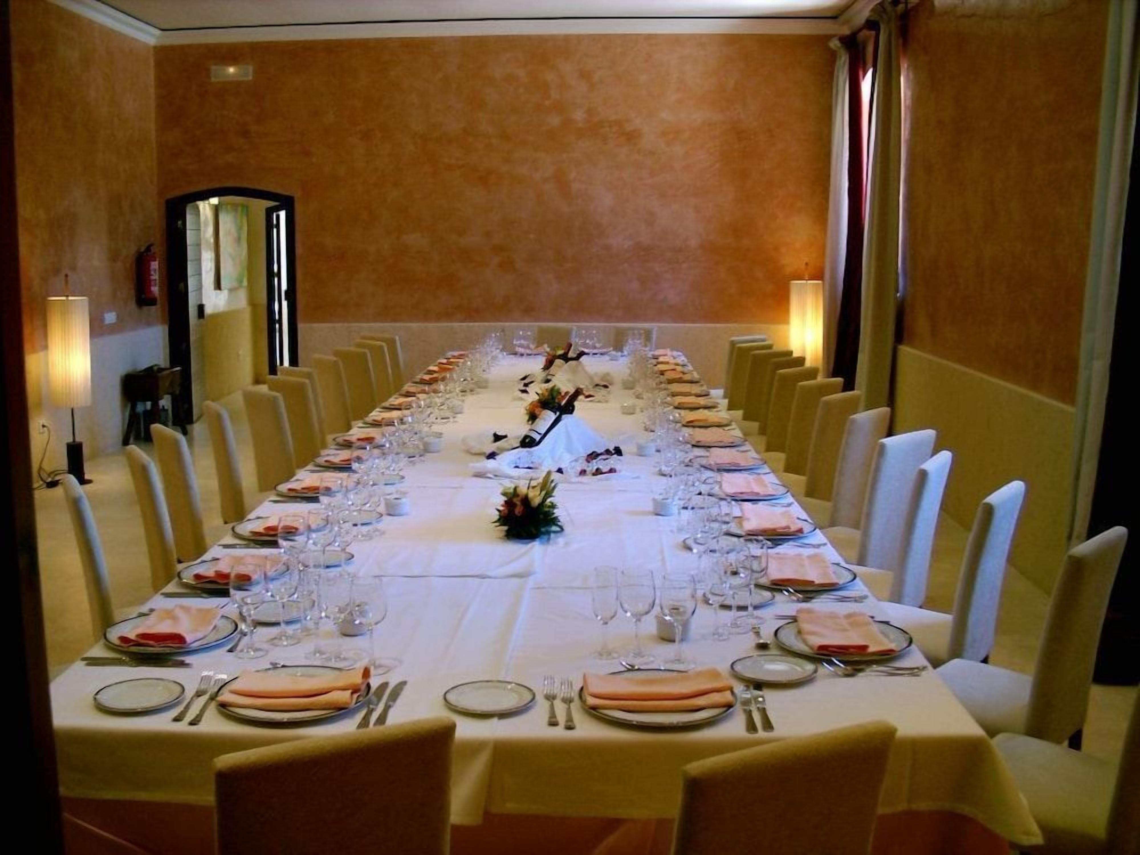 Una larga mesa de comedor dispuesta para una comida formal, con múltiples sillas, mantel blanco y una disposición simétrica de platos, copas y cubiertos.