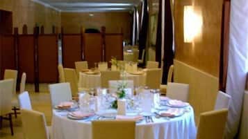 Une salle de banquet avec des tables rondes dressées pour le repas, des chaises et une longue table en arrière-plan.