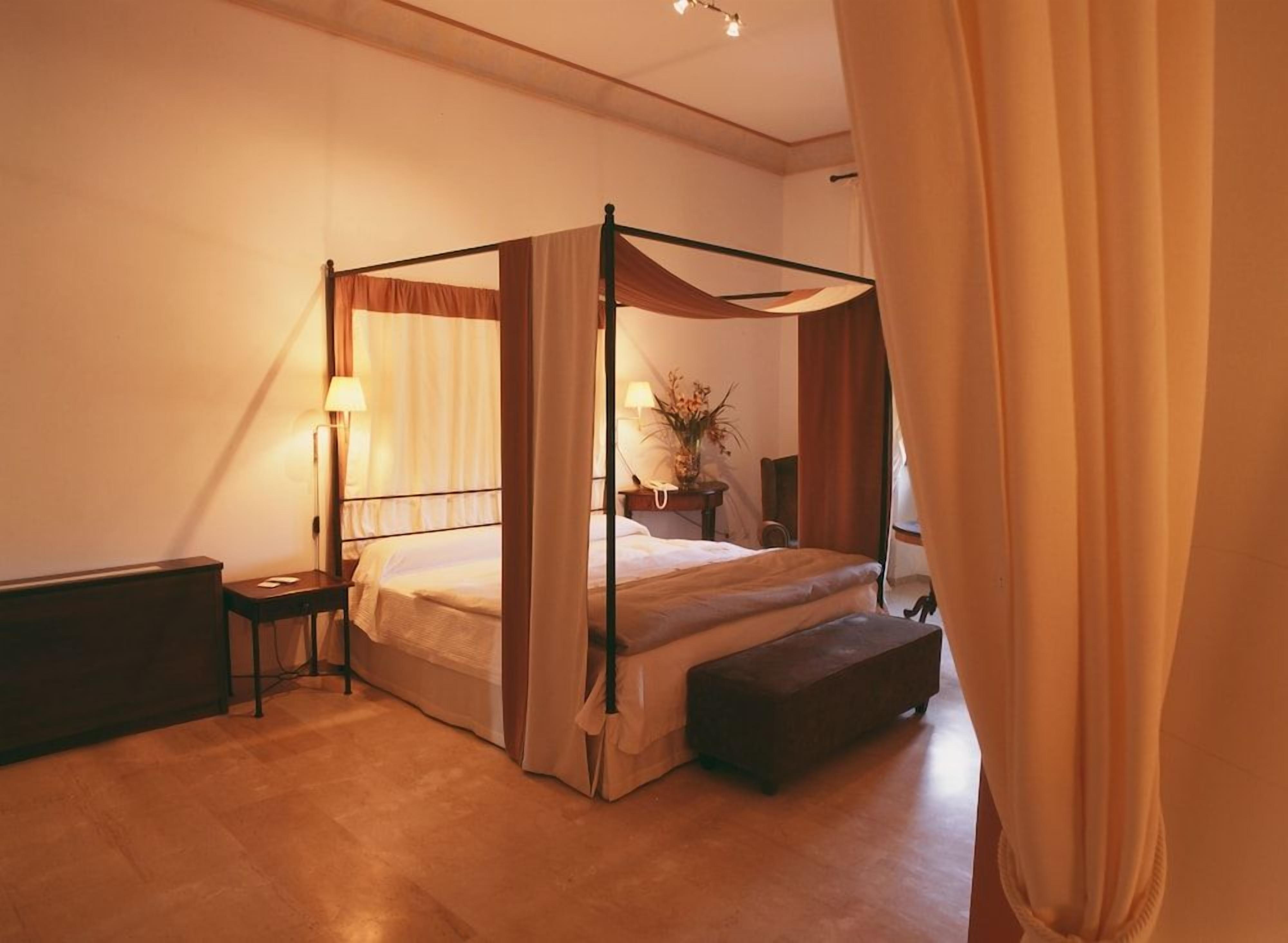 A four-poster bed with a bench, a bedside table, and a lamp in a hotel room.