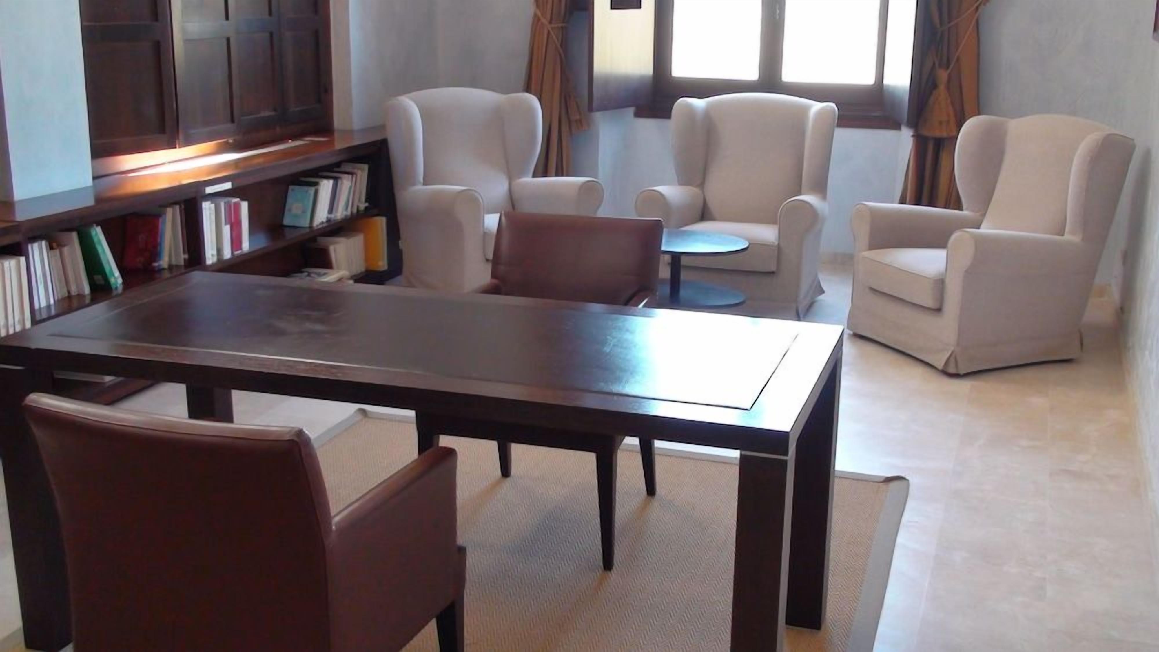 A modern living room with a dark wooden table, leather chairs, a bookshelf, and a large window with curtains.