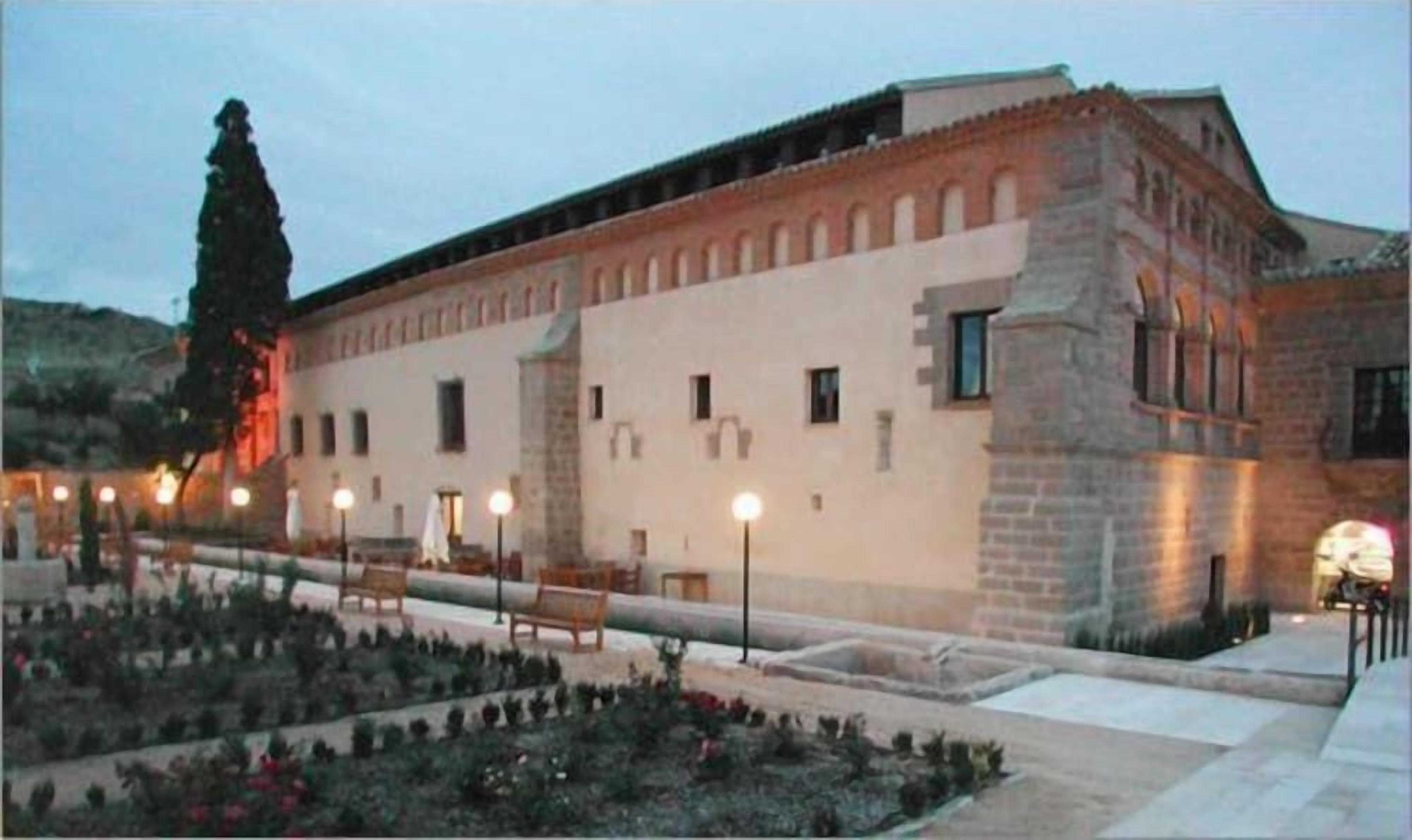 A building with a long, rectangular structure, a courtyard with benches, and a tall tree.