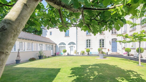 Een binnentuin met een gebouw, voorzien van grote ramen en een blauwe deur, omgeven door groen en bloempotten.