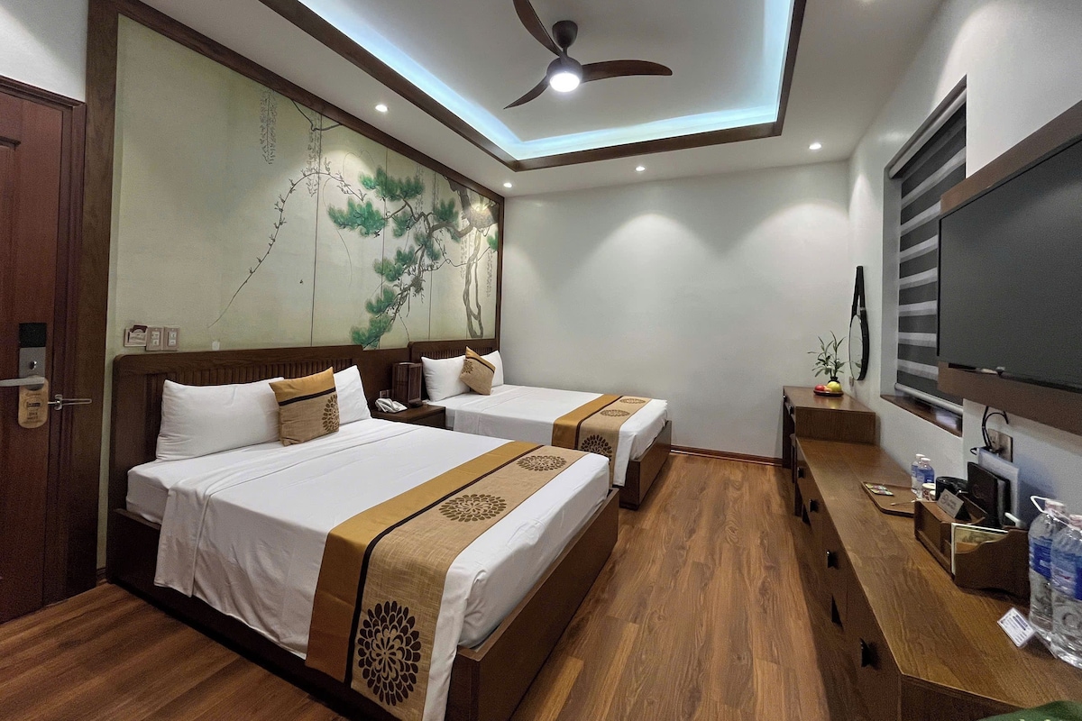 Habitación de hotel con dos camas, un ventilador de techo, un mural grande en la pared, un televisor y un escritorio de madera con una botella de agua.