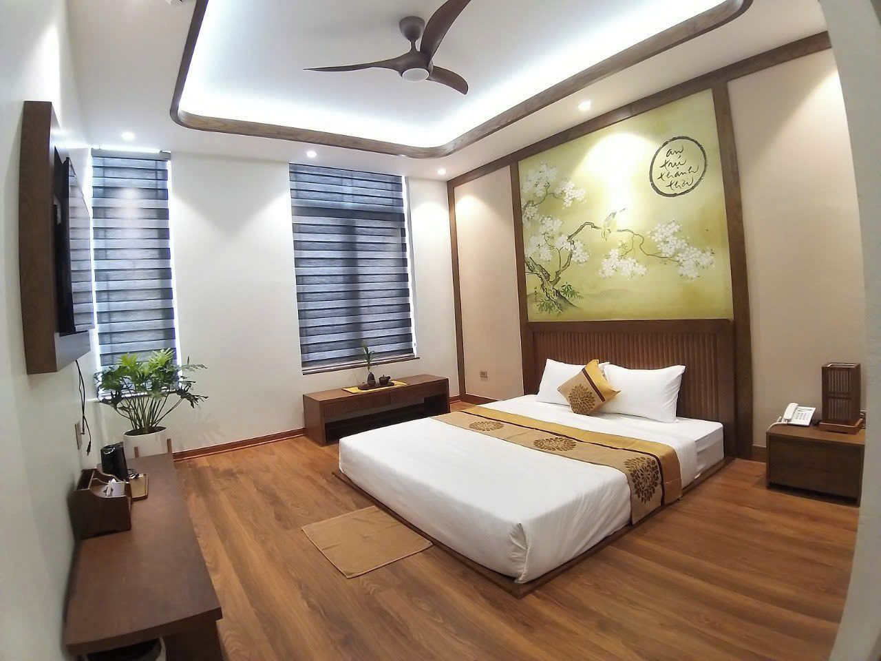 Habitación de hotel con cama, mesitas de noche, ventilador de techo, ventana con persianas, una obra de arte colgada en la pared y un teléfono en la mesita de noche.
