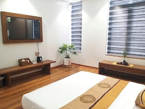 Una habitación de hotel con una cama grande, un banco de madera, una mesita con una tetera, una planta en maceta y un televisor montado en la pared.