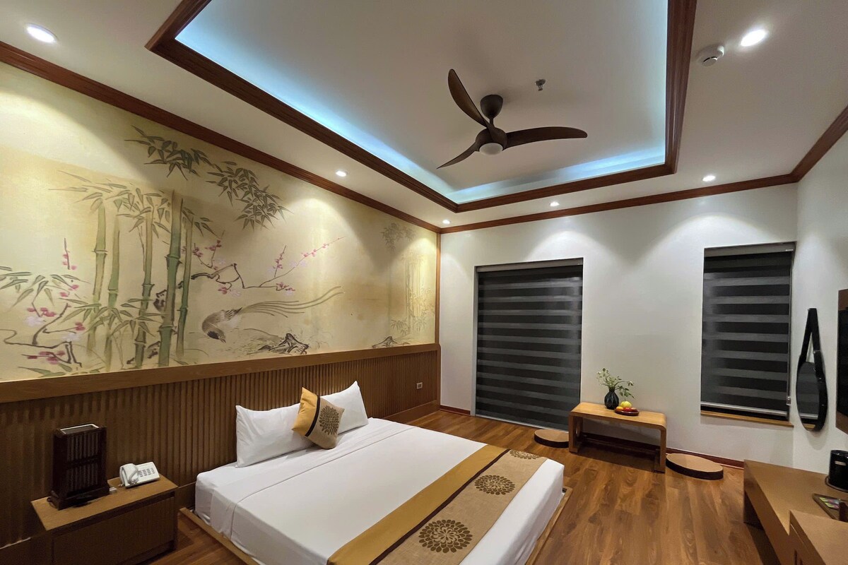 Habitación de hotel con una cama grande, suelo de madera, ventilador de techo, una pared con una pintura de bambú y una ventana con persiana.