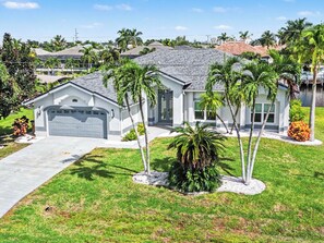 A modern two-story house with a garage, surrounded by well-maintained grass and palm trees. - Palms on 45th | Gulf Access, Heated Pool & Spa (Cape Coral)