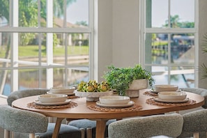 A round wooden dining table with white plates and bowls, surrounded by grey upholstered chairs, set against a backdrop of large windows overlooking a body of water. - Palms on 45th | Gulf Access, Heated Pool & Spa (Cape Coral)