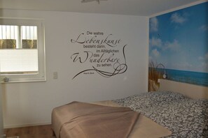 A bedroom with a bed, a window with blinds, a wall with a quote, and a painting of a beach. - Dinius Schützenstraße (Grömitz)