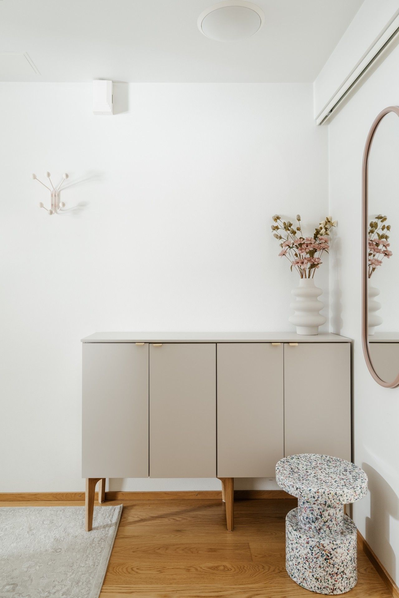 Une pièce au décor minimaliste, avec un meuble blanc, une petite table ronde, un miroir et un vase contenant des fleurs séchées.