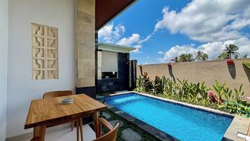 Zona de piscina con mesa y sillas de madera, rodeada de plantas tropicales y un cielo despejado.