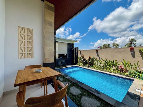 A pool area with a wooden table and chairs, surrounded by tropical plants and a clear sky.