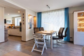 A modern dining area with a wooden table and chairs, a kitchen in the background, and a patterned curtain. - Holiday home with 2 bedrooms (Kellenhusen)