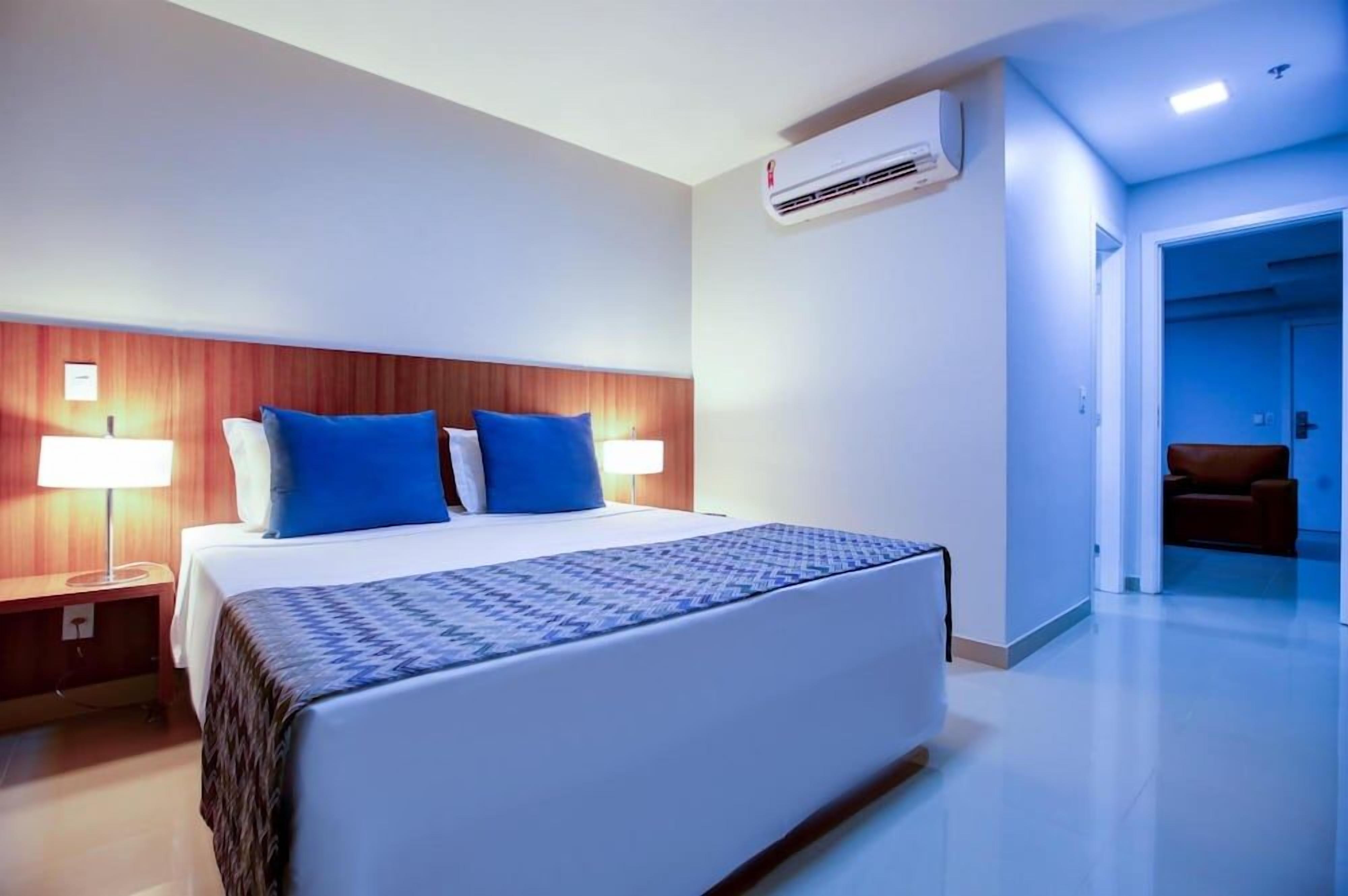 Quarto de hotel com uma cama, duas luminárias laterais, uma cabeceira de madeira, edredom estampado e ar condicionado.