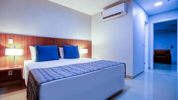 Quarto de hotel com uma cama, duas luminárias laterais, uma cabeceira de madeira, edredom estampado e ar condicionado.
