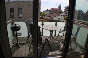 En balkong med ett runt bord och stolar, en grill och utsikt över kvarteret.