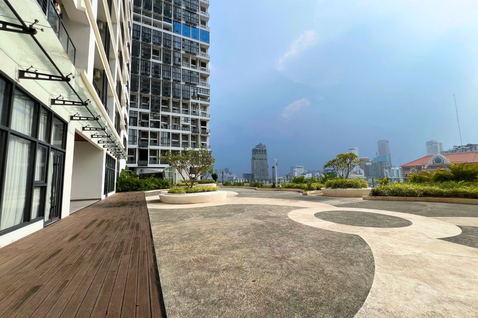 A rooftop terrace with a wooden deck, circular seating areas, and a view of a city skyline.