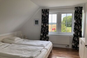 Ein Schlafzimmer mit zwei Betten, einem Fenster mit Vorhängen, einem Heizkörper und einem kleinen Nachttisch mit Lampe.