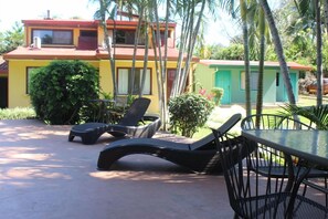 Un patio con sillones de descanso y una mesa, rodeado de vegetación y un edificio con techo rojo.