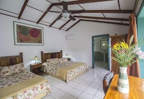 Un dormitorio con dos camas, un ventilador de techo, un cuadro en la pared, una mesita de noche con una lámpara y un florero con flores sobre una mesa de madera.