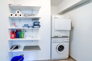 A laundry area with a stacked washer and dryer, shelves with cleaning supplies, and a blue bucket. - Holiday apartment with 2 bedrooms (Sankt Peter-Ording)