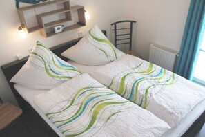 A bed with a white and green patterned bedspread, a wooden headboard, a bedside table, a wall-mounted shelf with a lamp, and a window with curtains. - Holiday apartment with 2 bedrooms (Wendisch Rietz)