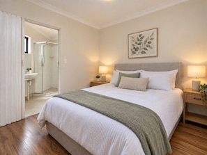 Quarto com cama, mesas de cabeceira, um criado-mudo, planta emoldurada e banheiro visível por uma porta aberta.