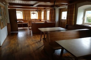 Ein geräumiges Zimmer mit Holzboden, einem großen Tisch und einer Bank. Es gibt Fenster mit Vorhängen, und eine Tür ist sichtbar.