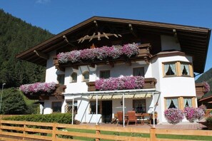 A two-story building with balconies adorned with flowers, a covered patio with seating, and a mountainous backdrop. - Country house - Praxmarer (Neustift im Stubaital)