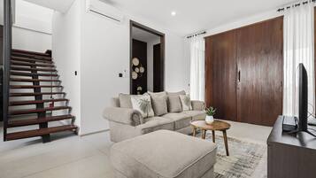 Una sala de estar moderna con sofá, reposapiés y una mesa de centro. Se ve una escalera de madera a la izquierda y una puerta de madera grande a la derecha.