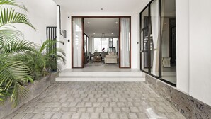 A modern interior with a hallway leading to a living area, featuring a brick floor, large windows, and indoor plants. - Allena Villa by Kozystay - Canggu (Badung)