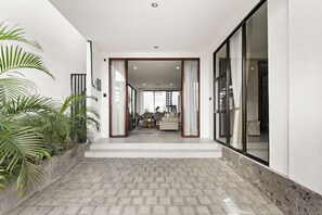 A modern interior with a hallway leading to a living area, featuring a brick floor, large windows, and indoor plants. - Allena Villa by Kozystay - Canggu (Badung)