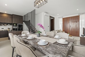 A modern kitchen with dark cabinets, a built-in oven, and a dining area with a marble table set for six. - Allena Villa by Kozystay - Canggu (Badung)