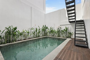 A rectangular swimming pool with clear water, surrounded by a white wall and a wooden deck. - Allena Villa by Kozystay - Canggu (Badung)