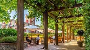 Ein Terrassenbereich im Freien mit Holzbögen, Tischen und Stühlen, umgeben von viel Grün und einem historischen Gebäude mit einem Glockenturm im Hintergrund.
