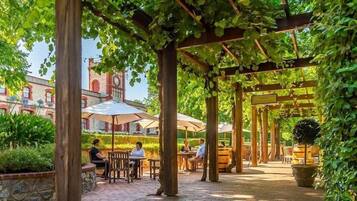 An outdoor patio area with wooden pergolas, tables, and chairs, surrounded by greenery and a historic building with a clock tower in the background.