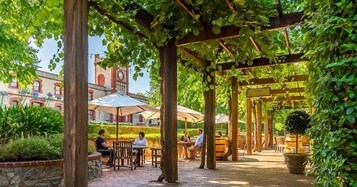 Un patio exterior con pérgolas de madera, mesas y sillas, rodeado de vegetación y un edificio histórico con una torre de reloj al fondo.