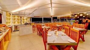 A spacious restaurant with multiple dining tables and chairs, a bar area, and a kitchen visible in the background. - Myspace Dhruva Inn (Bengaluru)