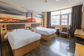 Hotellihuone, jossa on kaksi sänkyä, työpöytä, tuoli, pieni pöytä ja suuri ikkuna, jossa on verhot.