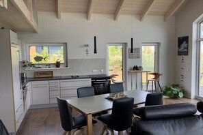 Une cuisine moderne avec des meubles blancs, une table à manger avec des chaises et une vue sur l’extérieur grâce à de grandes fenêtres.