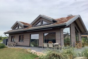 A modern two-story house with a sloped roof, large windows, and a patio area with outdoor furniture. - Painters' Corner Hohwacht (Hohwacht)