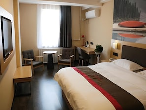 Hotellihuone, jossa on sänky, työpöytä, kaksi tuolia, pieni pöytä, televisio ja ikkuna, jossa on verhot.