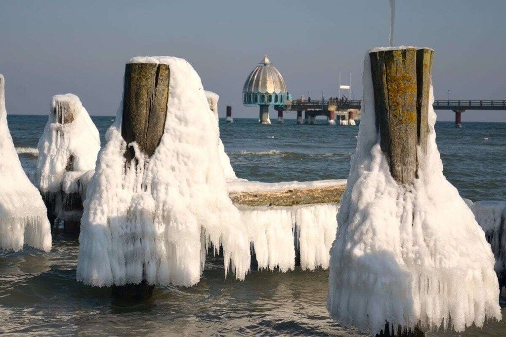 A pier with ice formations around the posts, a bridge, and a dome structure in the background.