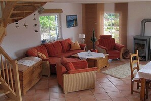 A cozy living room with a red sofa, a wooden chest of drawers, a wicker chair, and a fireplace. - Holiday home in Rügen with 2 bedrooms (Putgarten auf Rügen)