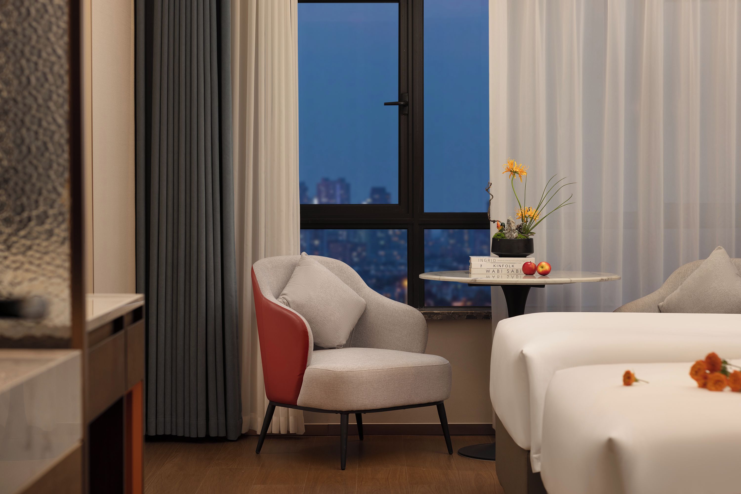 A hotel room with a bed, a chair, a small table with a vase of flowers, and a window with a city view.