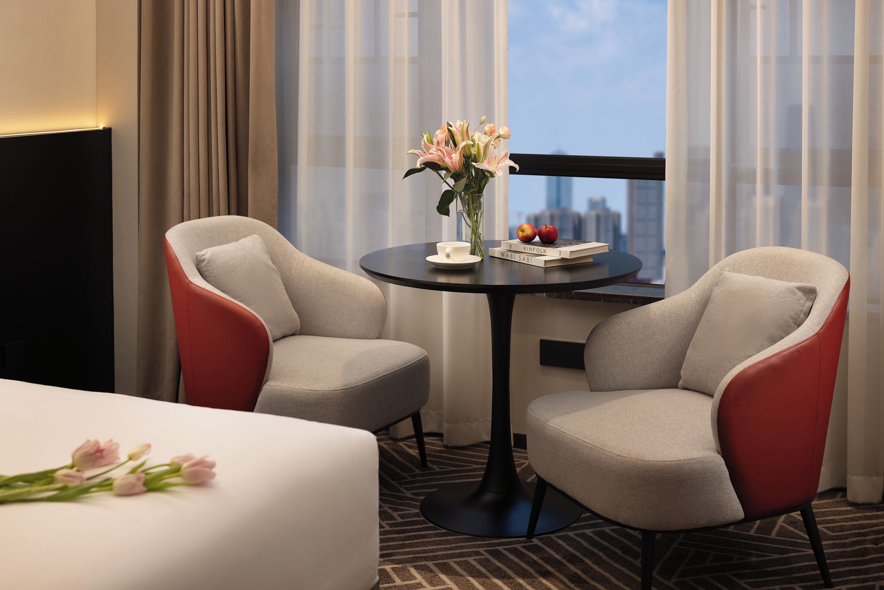 Habitación de hotel con dos sillones, una mesita redonda con un florero y vistas a un paisaje urbano a través de una ventana.