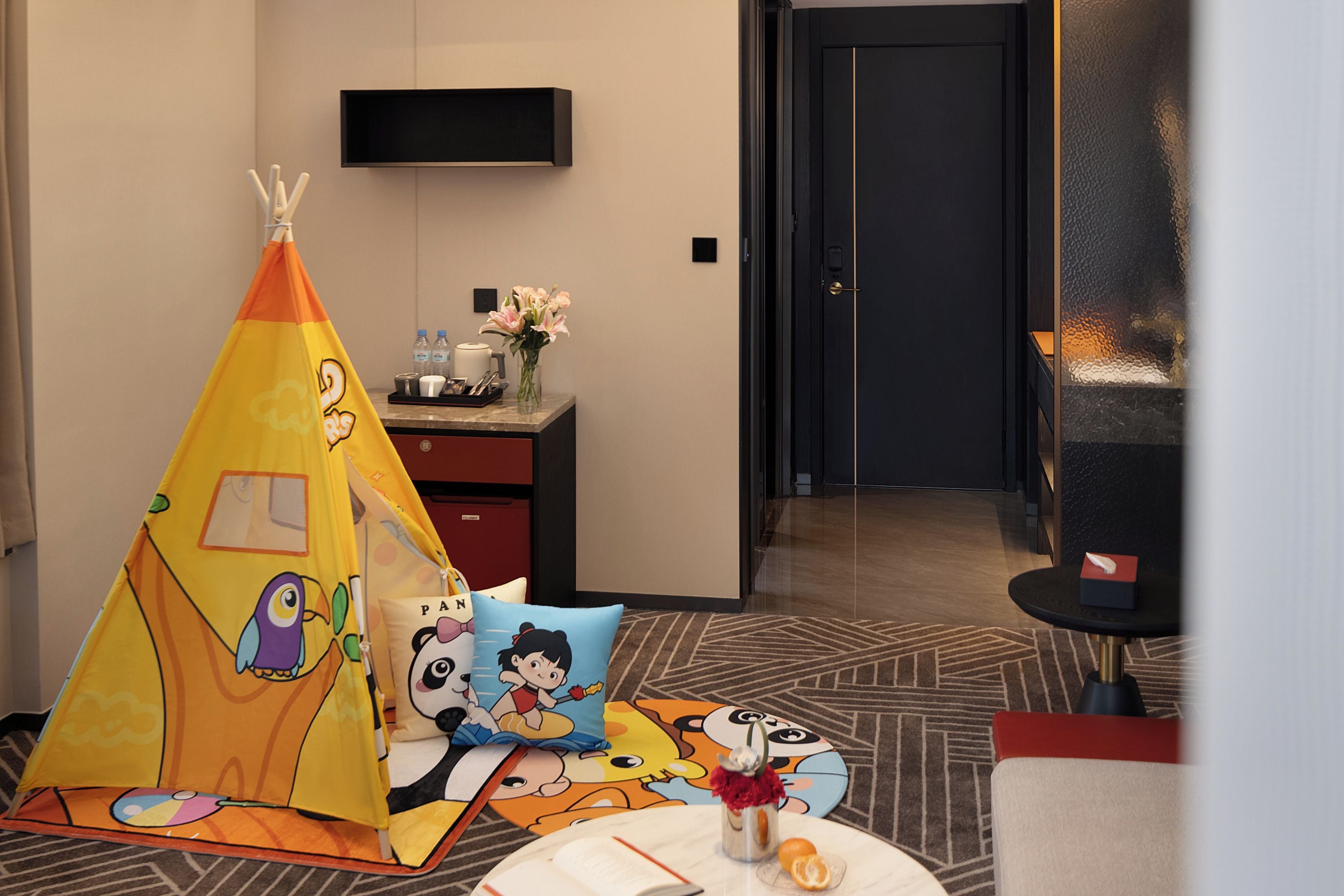 A hotel room with a children's play tent, a flat-screen TV mounted on the wall, a desk with a vase of flowers, and a door leading to another room.