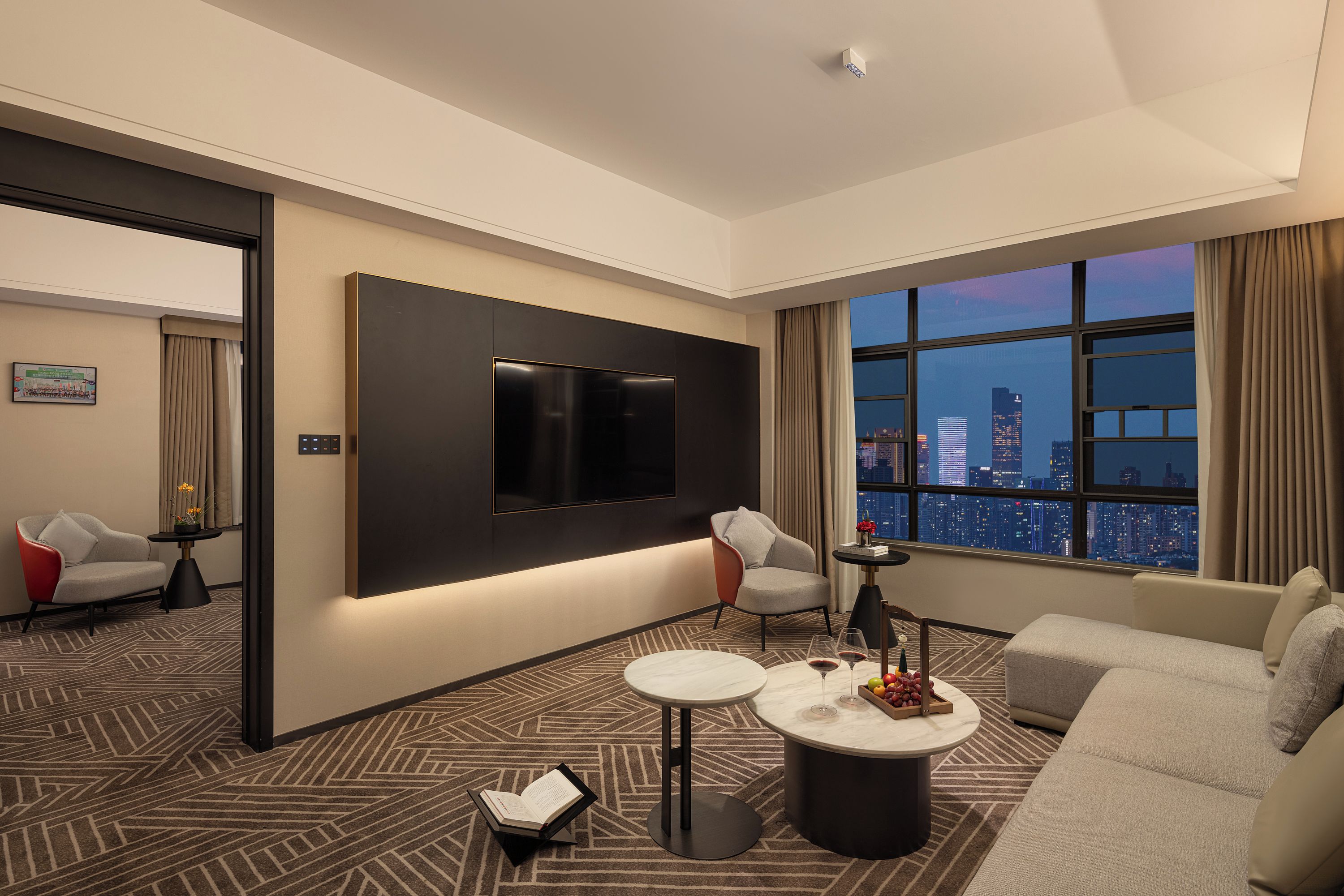 A modern hotel room with a large TV, a sofa, a coffee table, and a city view.