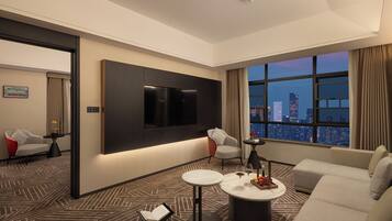 A modern hotel room with a large TV, a sofa, a coffee table, and a city view.