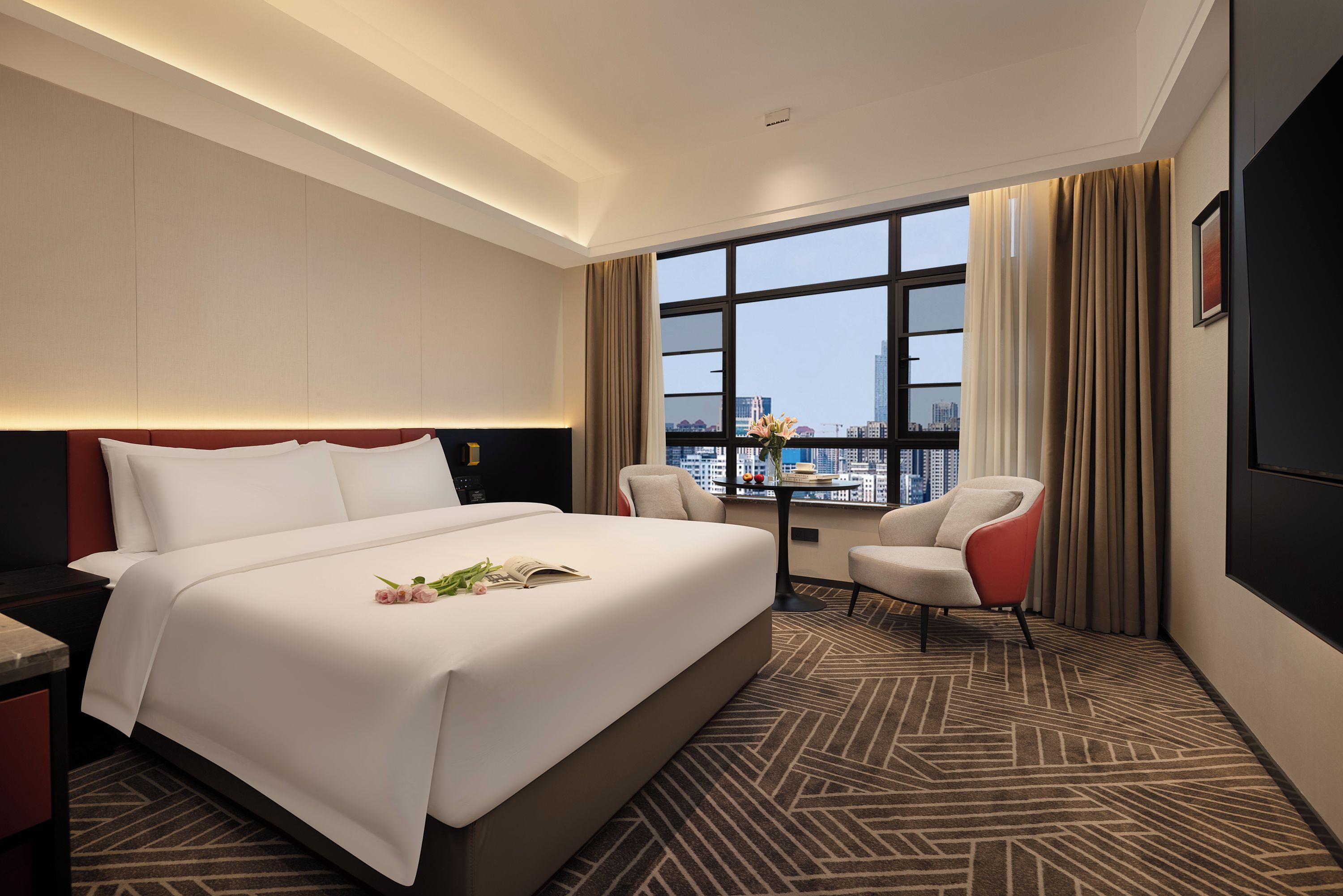 Habitación de hotel con una cama grande, un escritorio, una silla, un ventanal con vistas a la ciudad y una alfombra estampada.